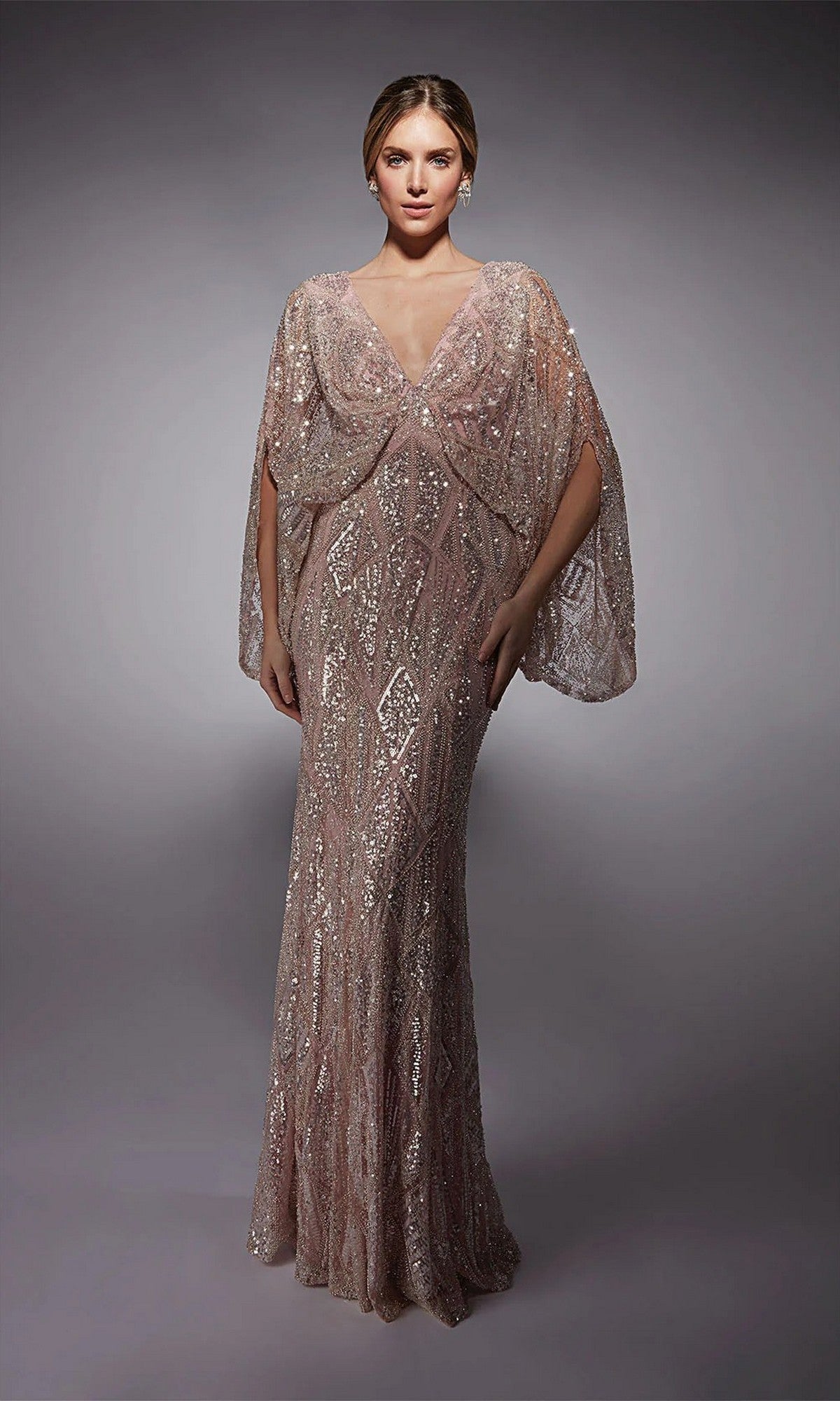 A woman in the Alyce 27756 long formal dress in cameo pink, featuring intricate geometric sequin patterns and a sheer cape-like overlay, stands against a plain gray background.